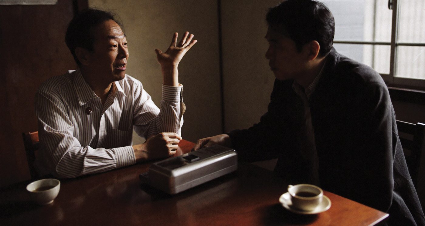 小さな事務所の机をはさんで、社長が話し、聞き手がメモを取らずに聞いている。机の上には録音機とお茶。